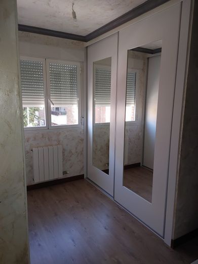 Habitación vacía con pisos de madera, puertas de armario con espejos y una ventana con persianas cerradas.