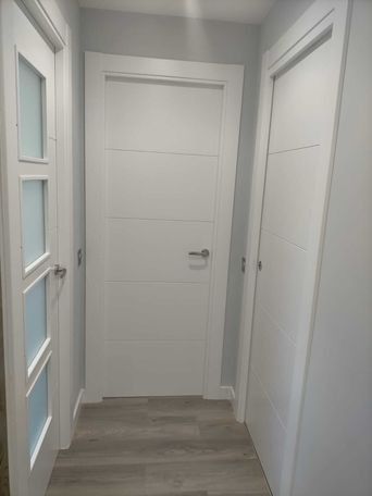 Puertas blancas en un pasillo con suelo gris; una puerta con paneles de cristal.