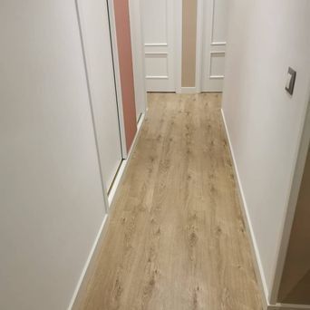 Pasillo estrecho con suelo de imitación madera clara, paredes blancas y una puerta cerrada al frente.