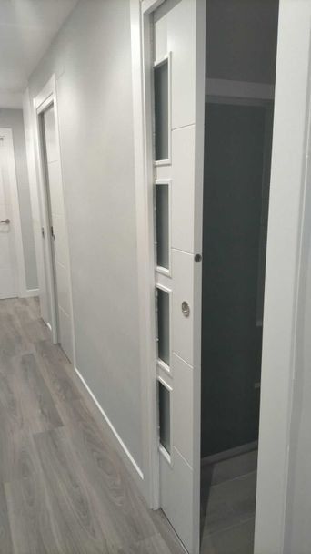 Un pasillo con una puerta corredera blanca parcialmente abierta y paneles de vidrio. Paredes gris claro y suelo imitación madera.