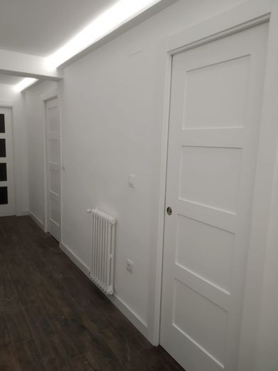 Pasillo blanco con puertas, radiador y suelo de madera oscura.