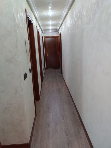 Pasillo estrecho con suelo de imitación madera y múltiples puertas de madera cerradas a los lados; paredes de color blanquecino.