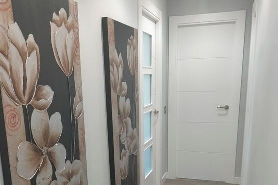 Un pasillo estrecho y blanco con dos cuadros florales a la izquierda y dos puertas blancas a la derecha.
