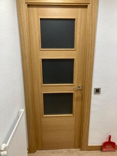 Puerta de madera con tres paneles de vidrio esmerilado, junto a una pared blanca y un pequeño recogedor de basura rojo.