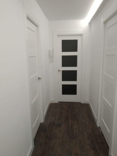 Pasillo estrecho con paredes blancas, piso de madera oscura y tres puertas blancas; una puerta tiene cuatro paneles de vidrio esmerilado.