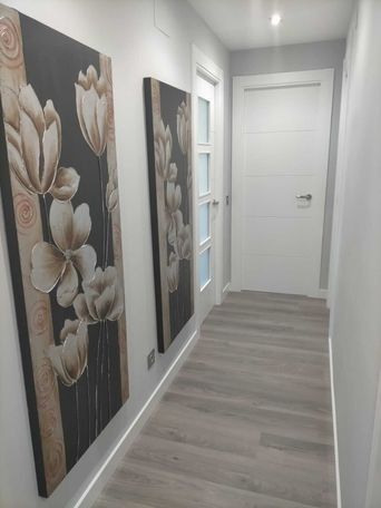 Pasillo estrecho con suelo de madera gris y paredes blancas, dos cuadros florales.