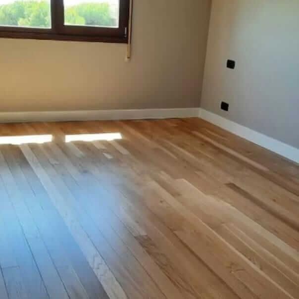 Habitación vacía con piso de madera, paredes beige y una ventana que deja entrar la luz del sol.