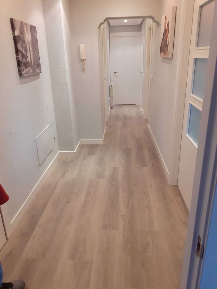 Pasillo estrecho con suelo de imitación madera clara, paredes blancas y una puerta cerrada al final.