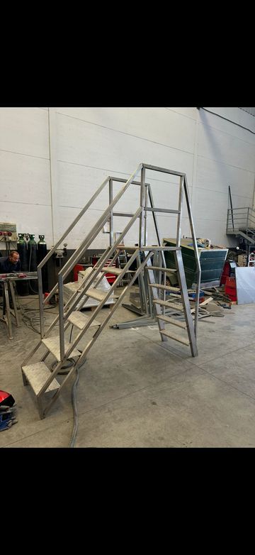 Plataforma metálica con escalera en un almacén, con pasamanos y una escalera de mano con ruedas delante.