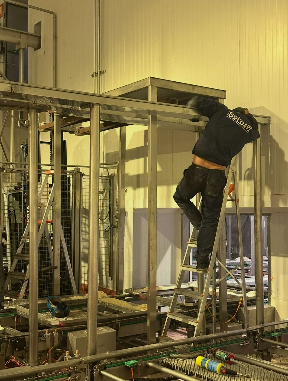 Un trabajador, subido a una escalera, repara maquinaria industrial dentro de una fábrica.