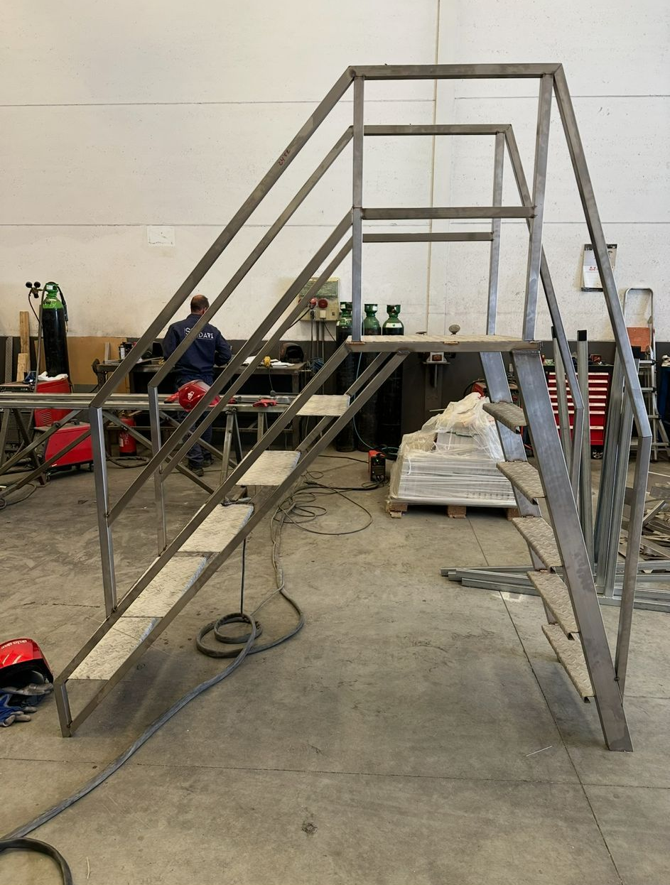 Escalera metálica con estructura en forma de A en construcción en un taller, con escalones sin terminar y herramientas cerca.