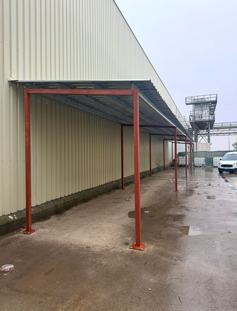 Estructura metálica roja tipo marquesina junto a un edificio industrial beige en un terreno de hormigón