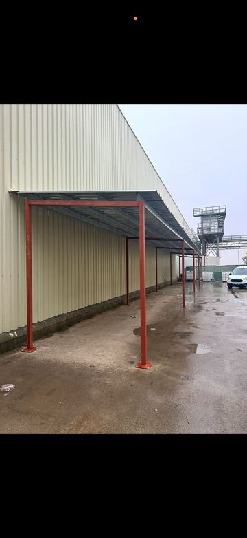 Un toldo metálico rojo se alza junto a un edificio industrial de color beige en un terreno de grava 