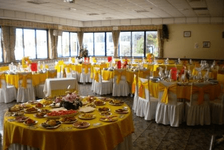 Salão de banquetes com toalhas de mesa amarelas, cadeiras brancas e mesas repletas de comida; as janelas oferecem uma vista.