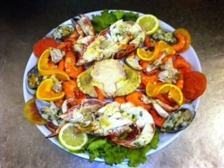 Prato de frutos do mar com lagosta, caranguejo, camarão, mexilhões, fatias de laranja e gomos de limão em um prato branco.