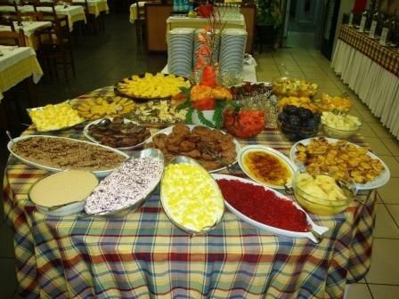 Mesa de buffet repleta de diversos pratos: saladas, queijos e pratos principais. Toalha de mesa xadrez.