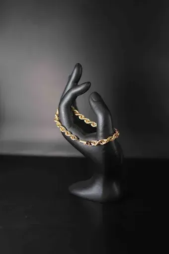 Mano de maniquí negra sosteniendo una pulsera de cadena dorada sobre un fondo oscuro.