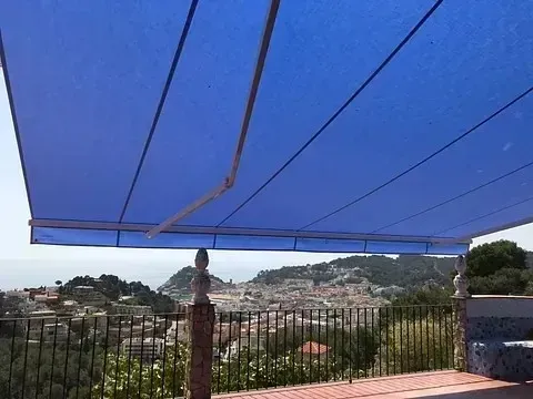 Un toldo azul da sombra a una terraza con vistas a un pueblo en la ladera de una colina y al mar a lo lejos.