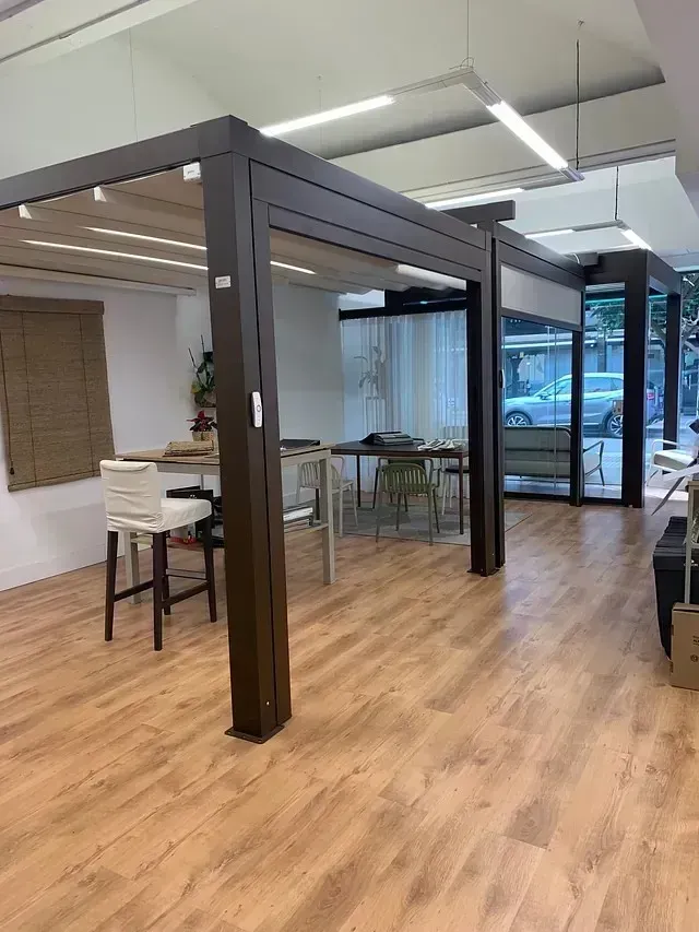 Una luminosa sala de exposiciones interior muestra modernas estructuras de pérgola de color marrón sobre mesas.