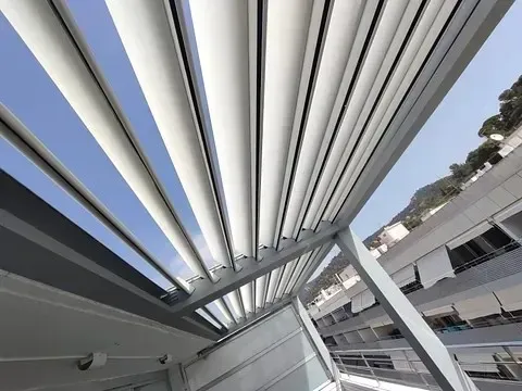 Vista desde un ángulo bajo de una pérgola moderna con lamas blancas ajustables, parcialmente abiertas.
