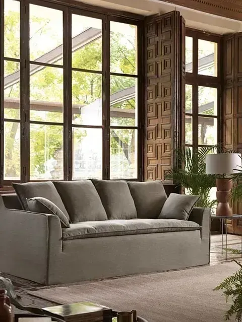 Un sofá beige con varios cojines descansa sobre una alfombra en una habitación con ventanas altas con marcos de madera.