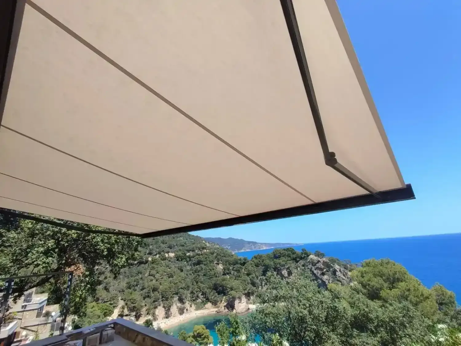 Un toldo beige se extiende sobre un balcón con vistas a una costa exuberante y montañosa.