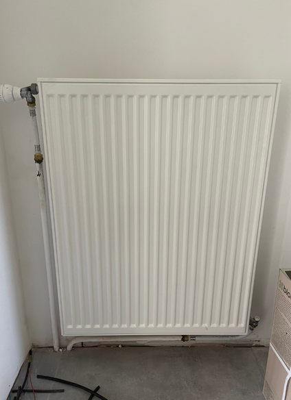 Radiateur mural blanc à barres verticales, raccordé à des tuyaux. Placé contre un mur blanc uni, dans une pièce.