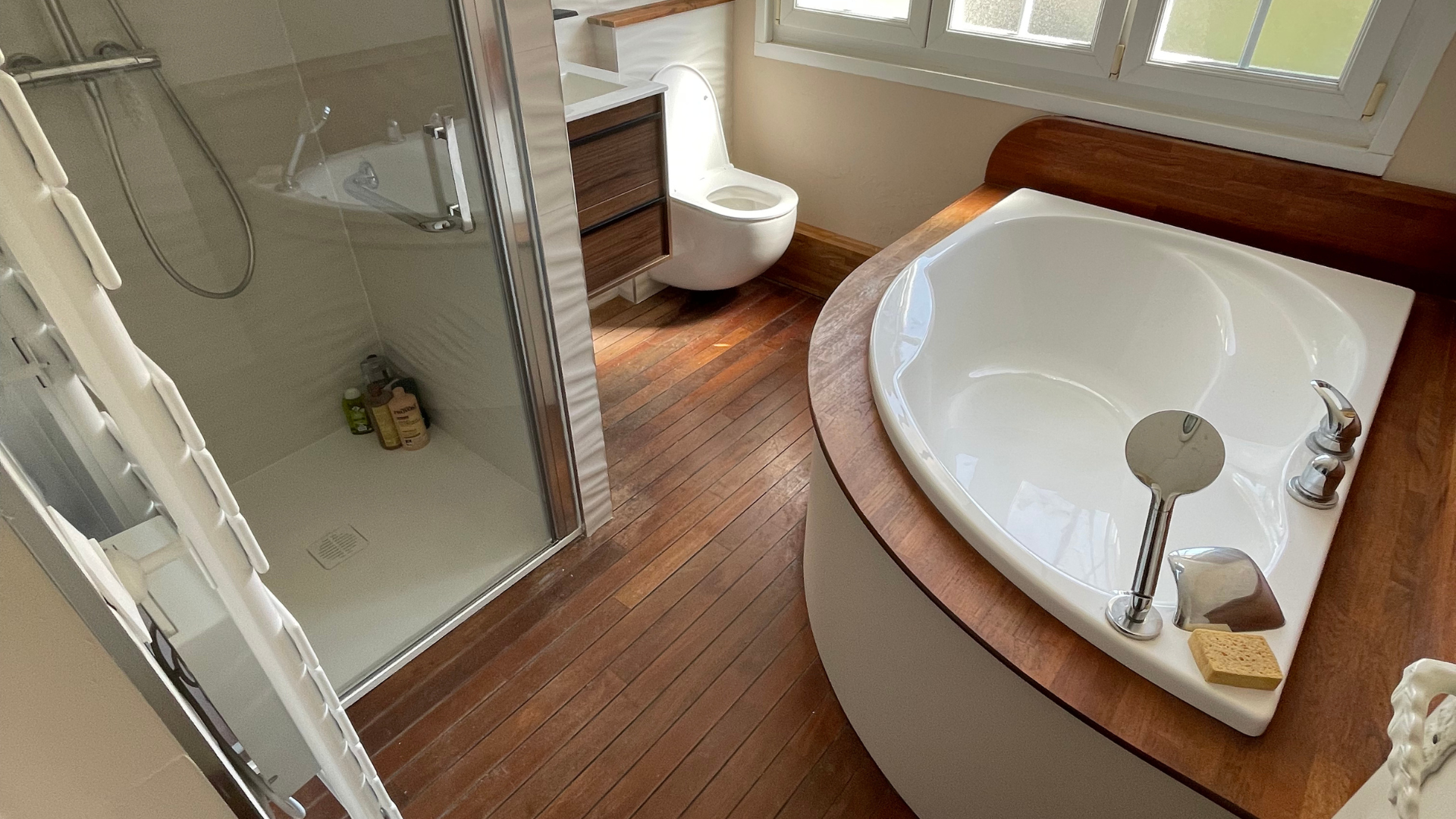 Salle de bain avec parquet, baignoire d'angle, douche, toilettes et vanité.