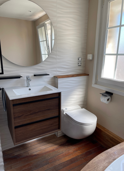 Salle de bain avec meuble-lavabo en bois et toilettes. Miroir ovale, lavabo blanc et mur texturé.