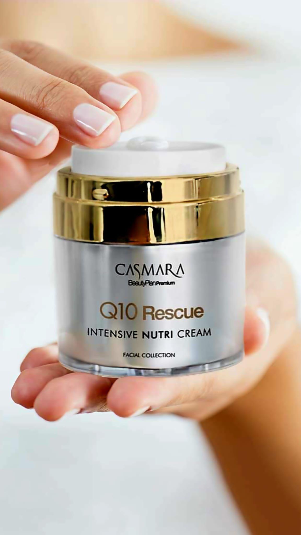 Una mano sosteniendo un frasco de crema Q10 Rescue de plata, con una tapa dorada, contra un fondo borroso.
