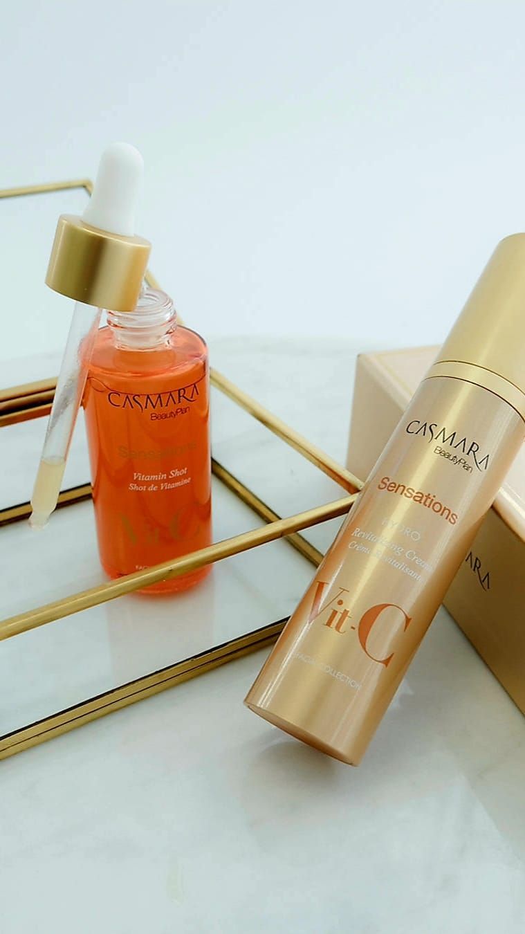 Dos frascos dorados para el cuidado de la piel, uno de ellos con gotero, sobre una bandeja de vidrio sobre un fondo blanco.