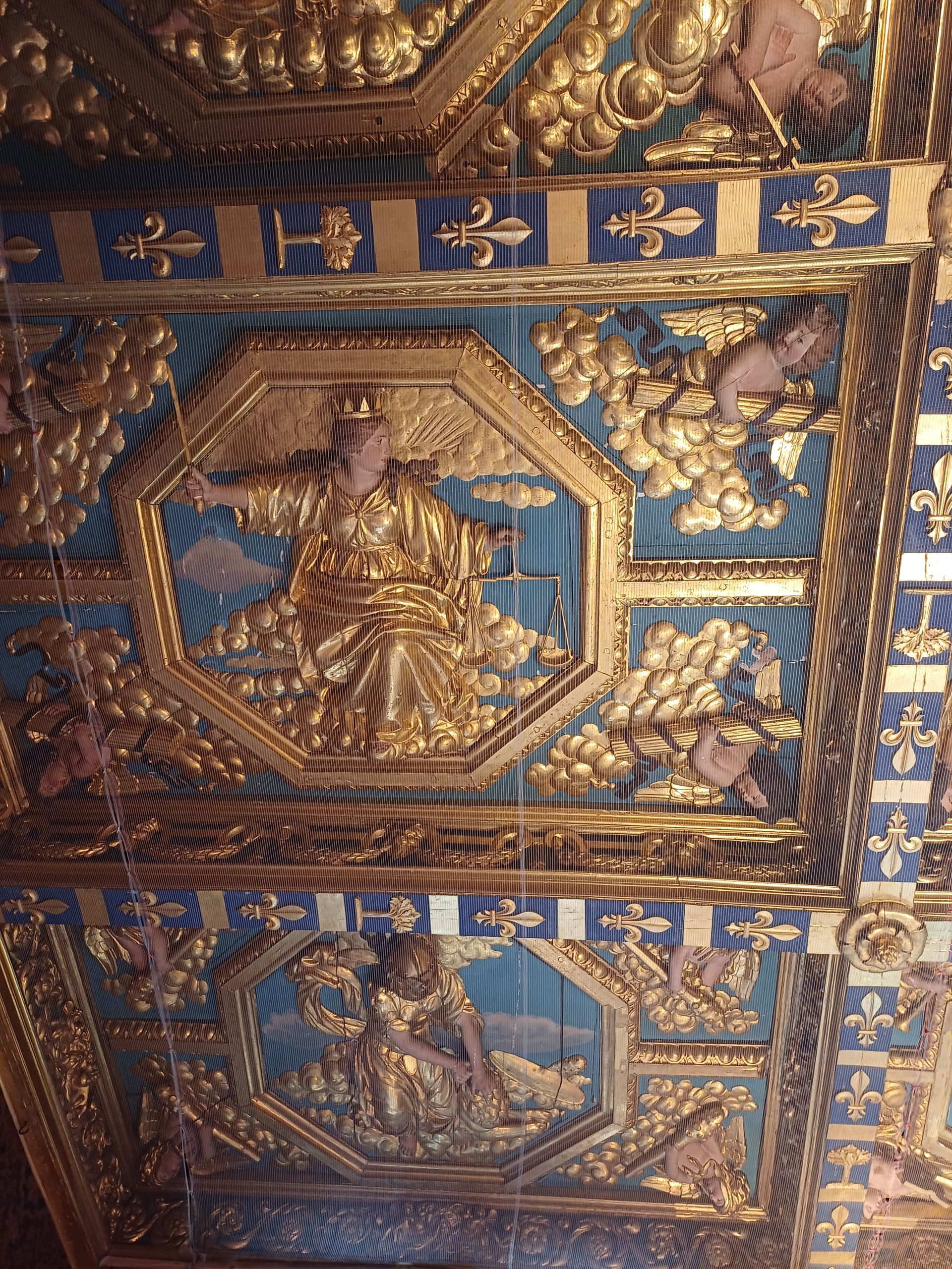 Des figures ornementales en bas-relief doré dans des cadres octogonaux sur un plafond bleu orné de fleurs de lys.
