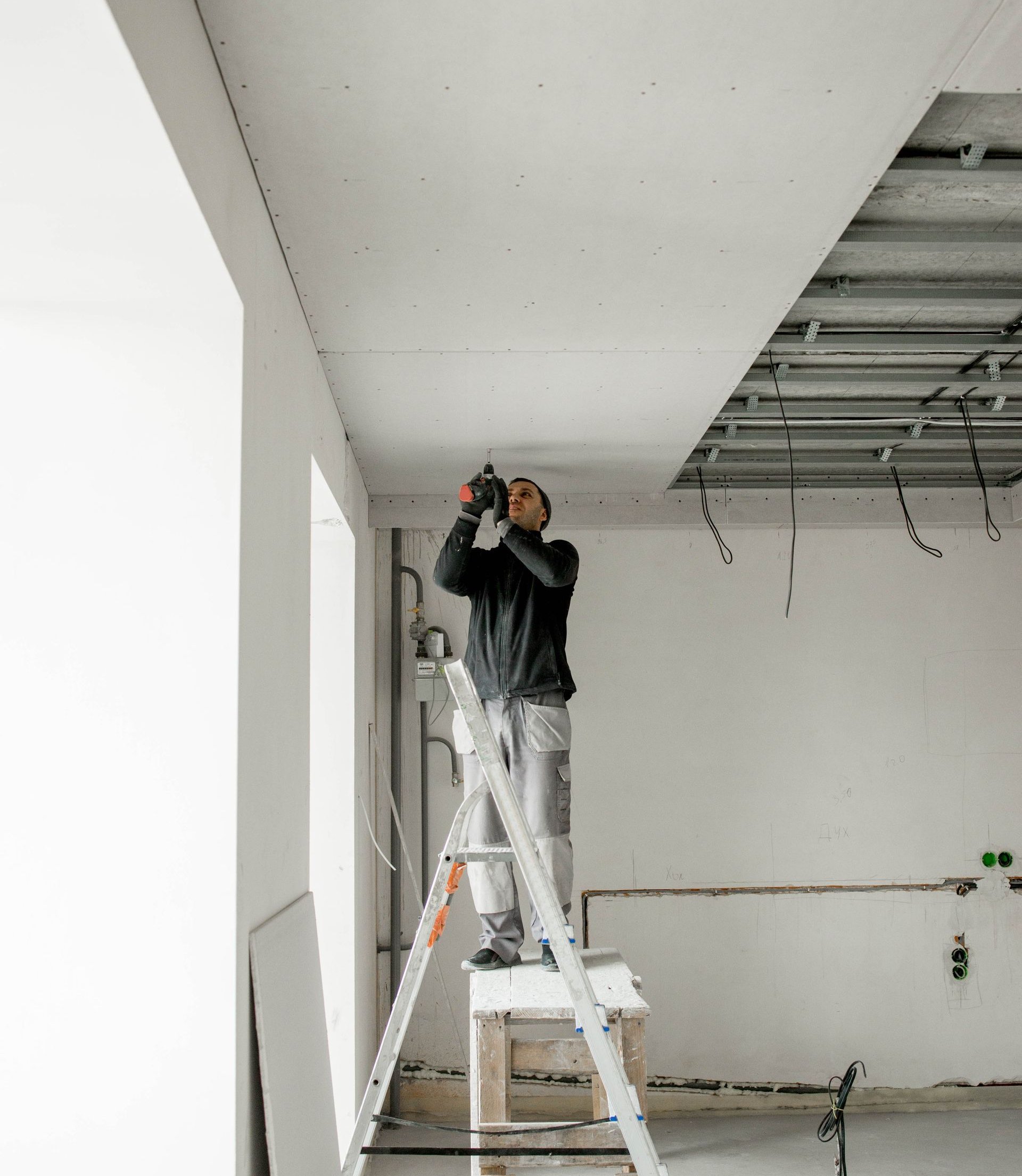 Un homme sur un escabeau pose des plaques de plâtre au plafond. Scène de chantier intérieur.
