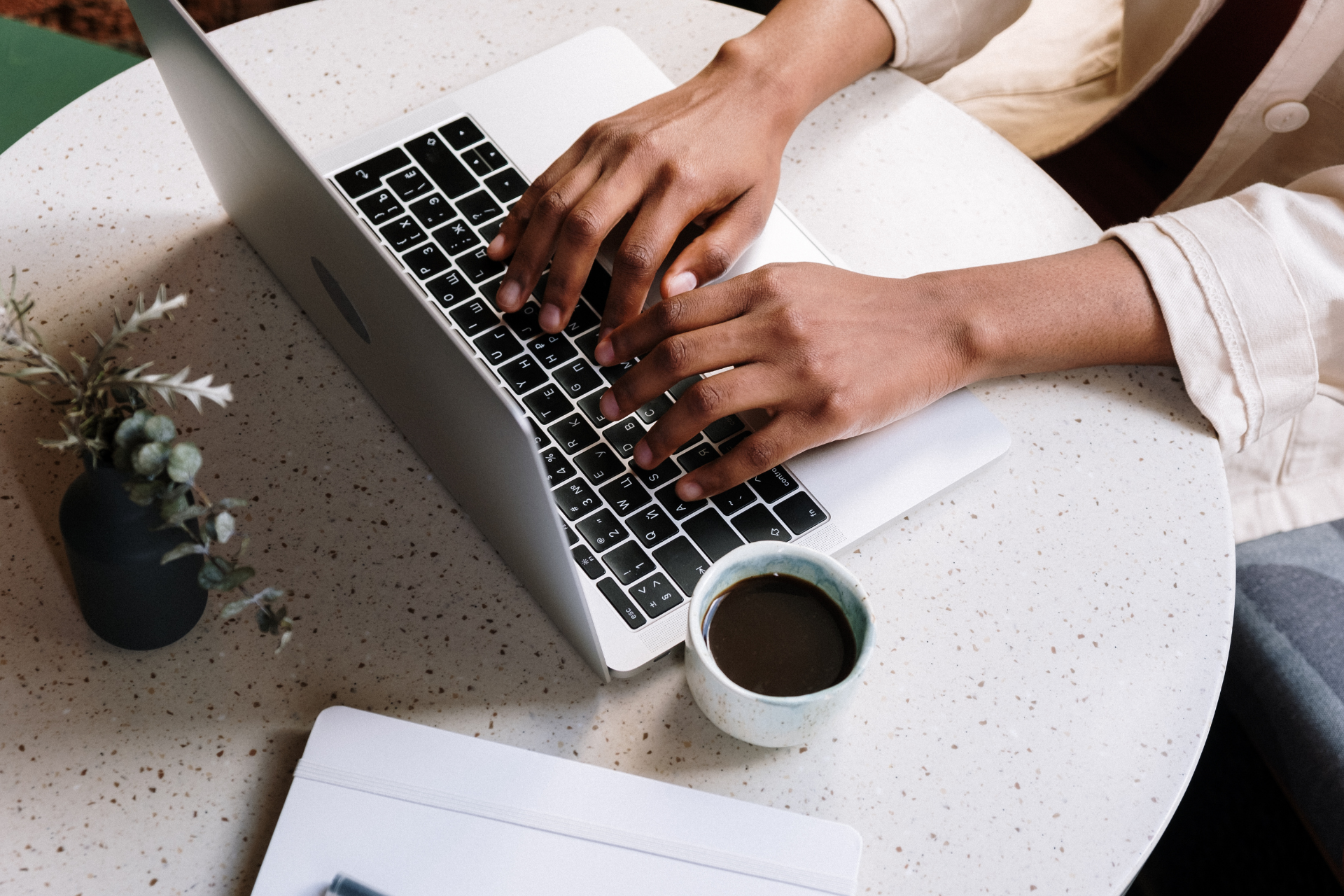 Manos escribiendo en una computadora portátil en una mesa redonda con una taza de café y una pequeña planta.