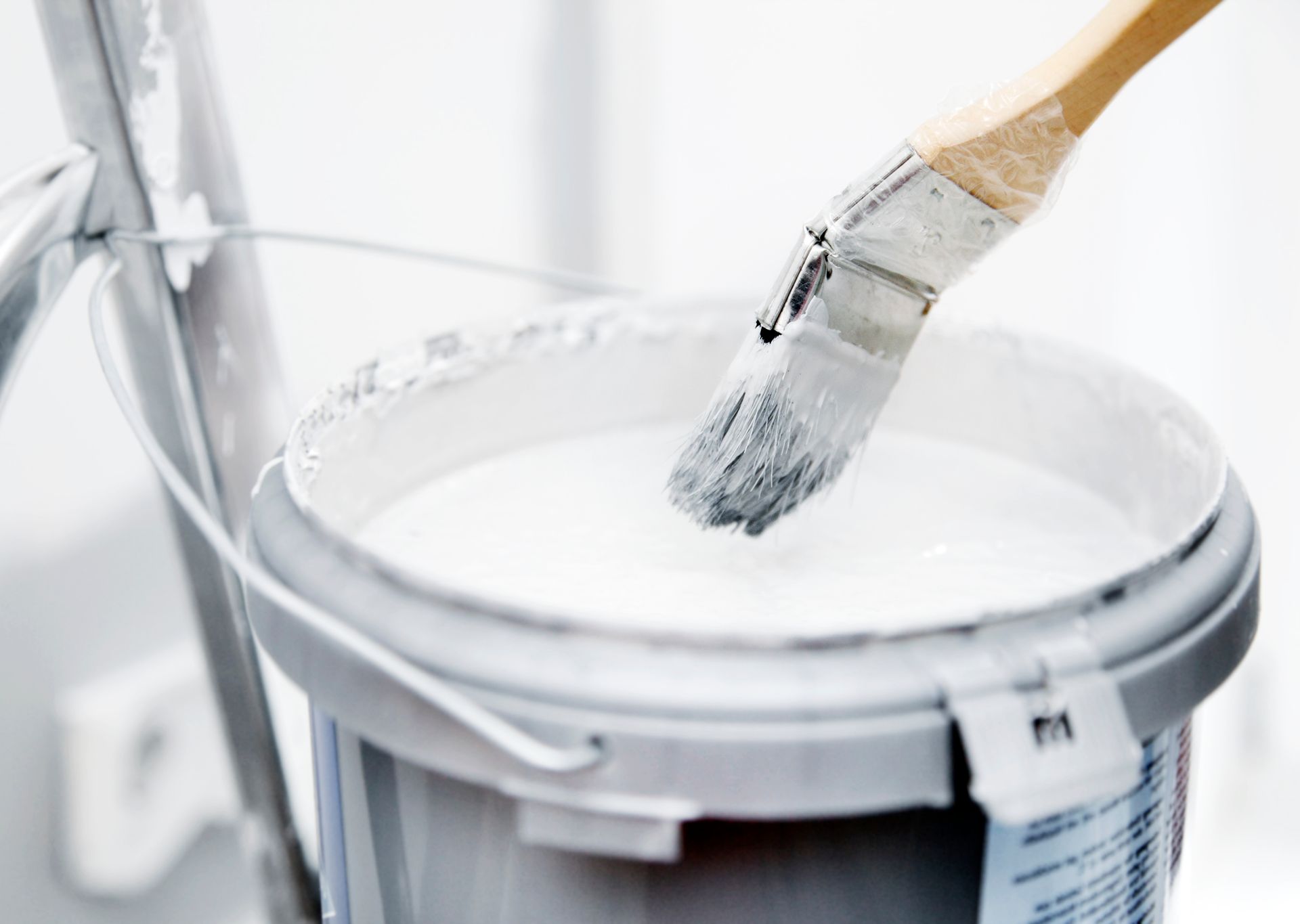 Ein Pinsel, der in einen Eimer weisser Farbe getaucht ist, steht neben einer Leiter.