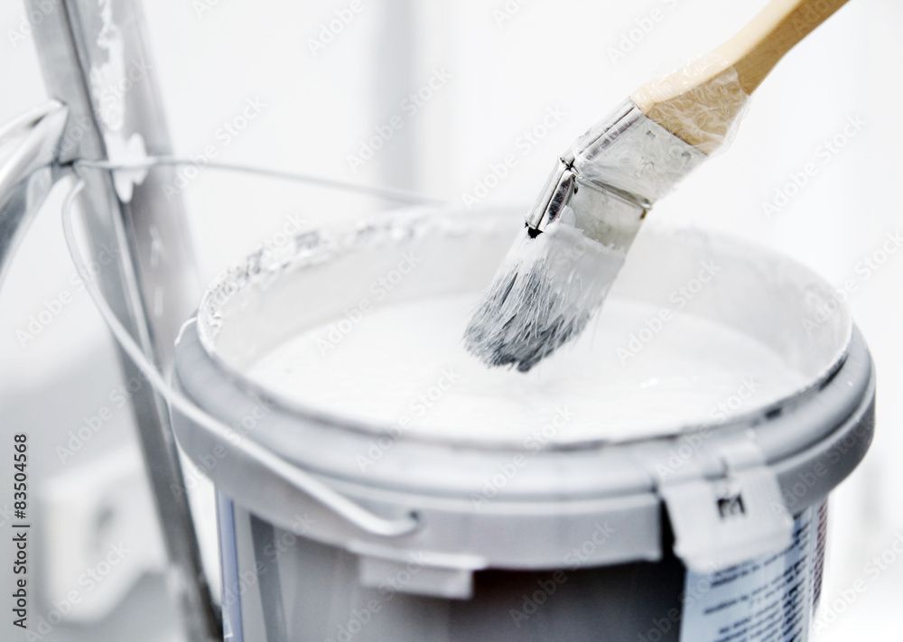 Ein Pinsel, der in einen Eimer weisser Farbe getaucht ist, steht neben einer Leiter.