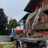 Une excavatrice sur un camion plateau devant un bâtiment en bois, avec une voiture au premier plan.