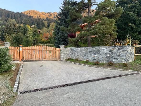 Entrée par un portail en bois et un mur de pierre, avec une allée pavée, dans un cadre montagneux.