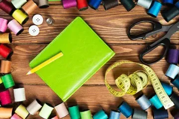 Un cuaderno verde está rodeado de carretes de hilo, tijeras, botones y una cinta métrica.