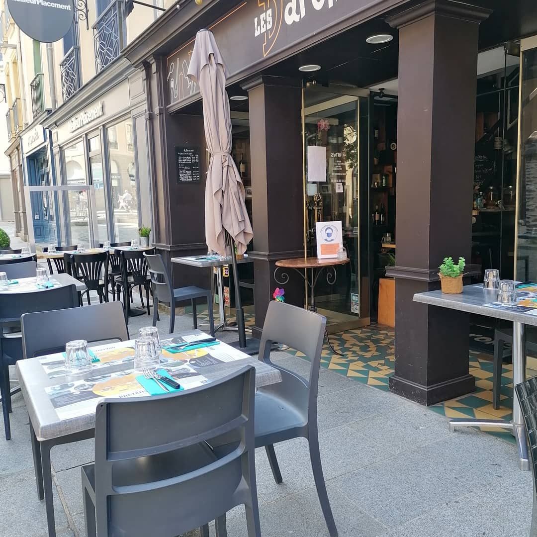 Restaurant en plein air avec tables et chaises installées sur le trottoir. L'entrée est ouverte et ombragée par un parasol partiellement fermé.