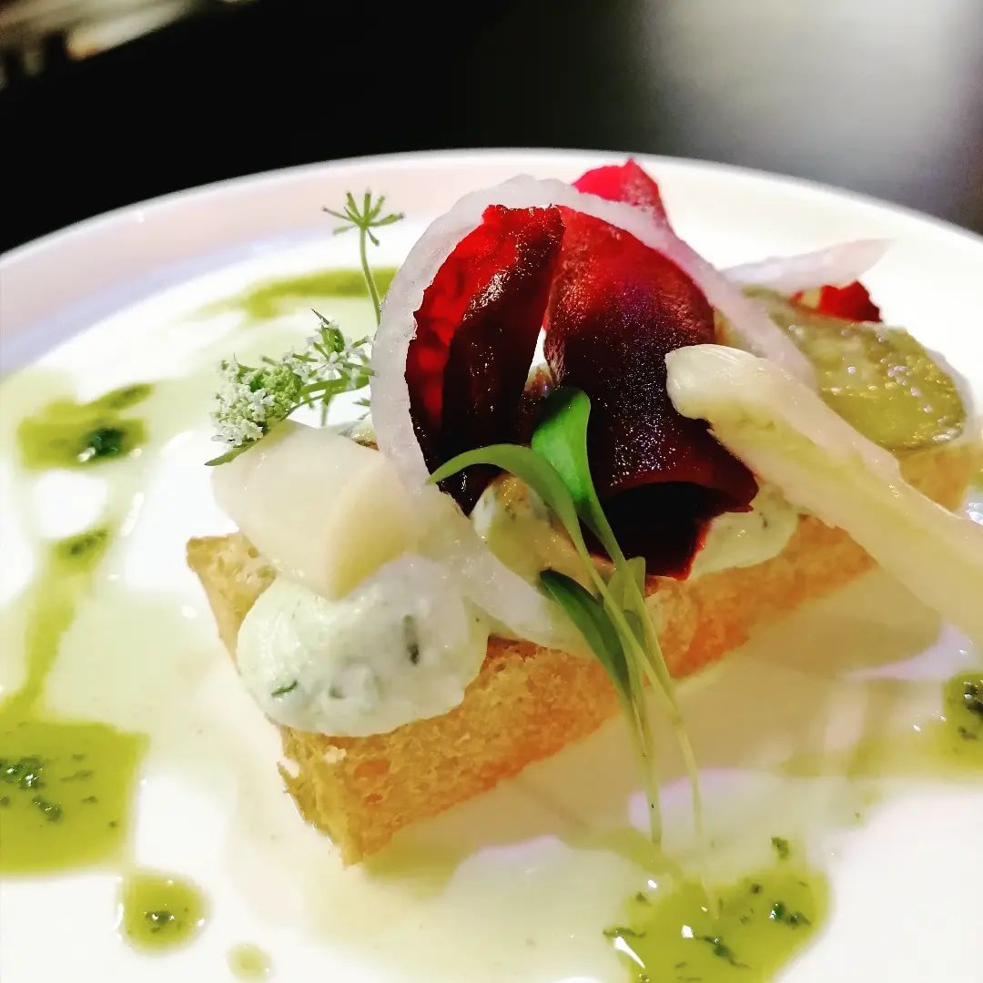 Entrée sur une assiette blanche : toast garni de betterave, d'oignon, d'asperges, d'herbes et de sauce verte.