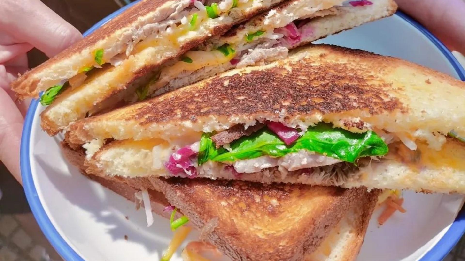 Des sandwichs grillés, coupés en deux, empilés dans un bol blanc à bord bleu, garnis de viande, de fromage et de légumes.