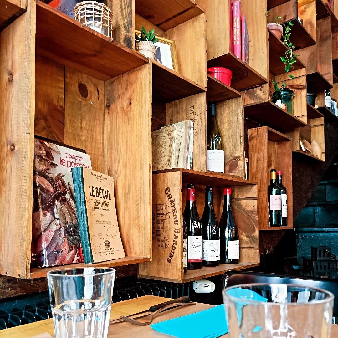 Étagères en bois avec livres, bouteilles et plantes dans un restaurant. Verres sur la table.