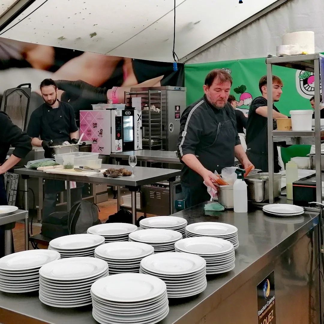Des chefs préparent des repas dans un stand professionnel ; des piles d'assiettes au premier plan.