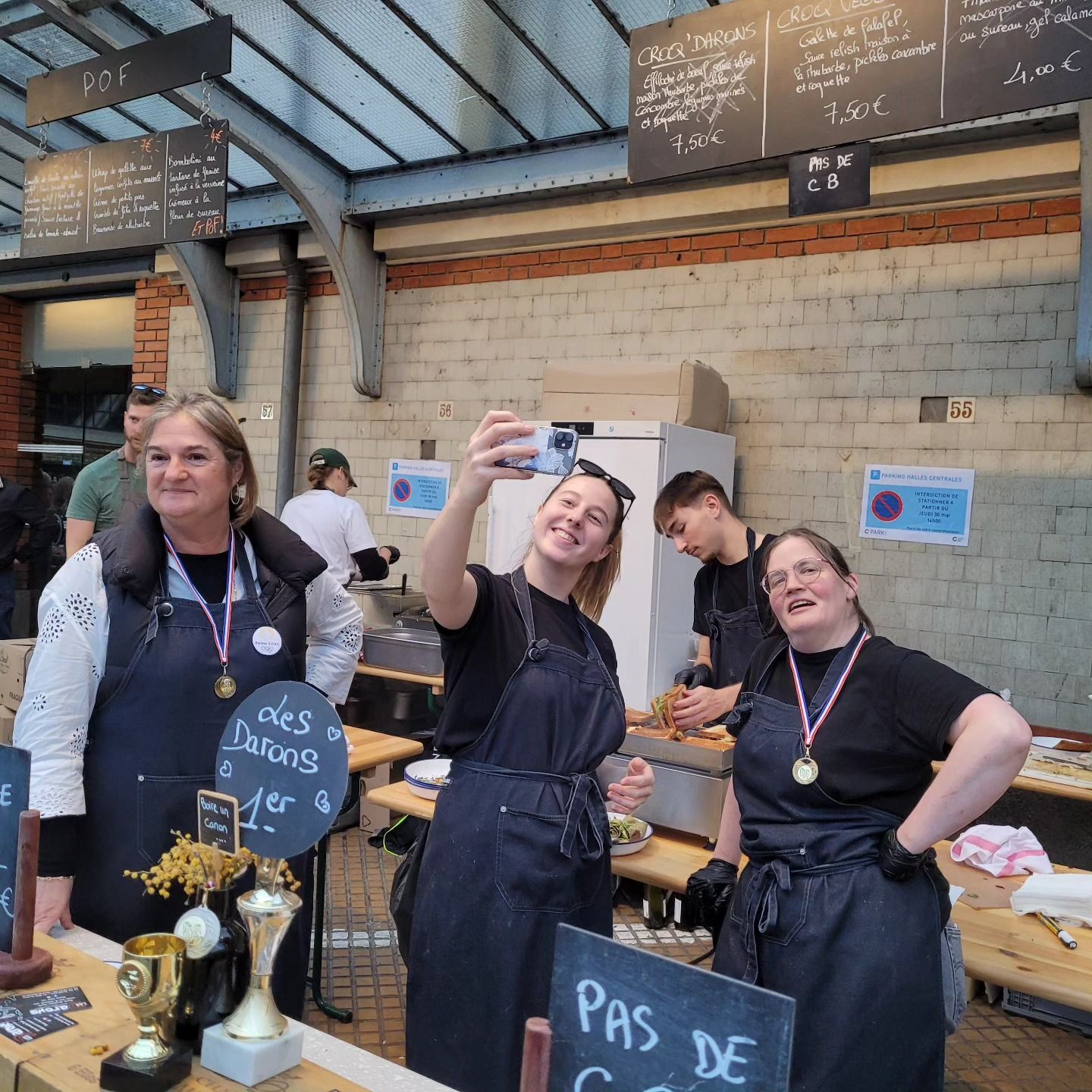 Quatre personnes portant des tabliers sur un étal de marché ; l’une prend un selfie, les autres sourient. Des médailles sont exposées.