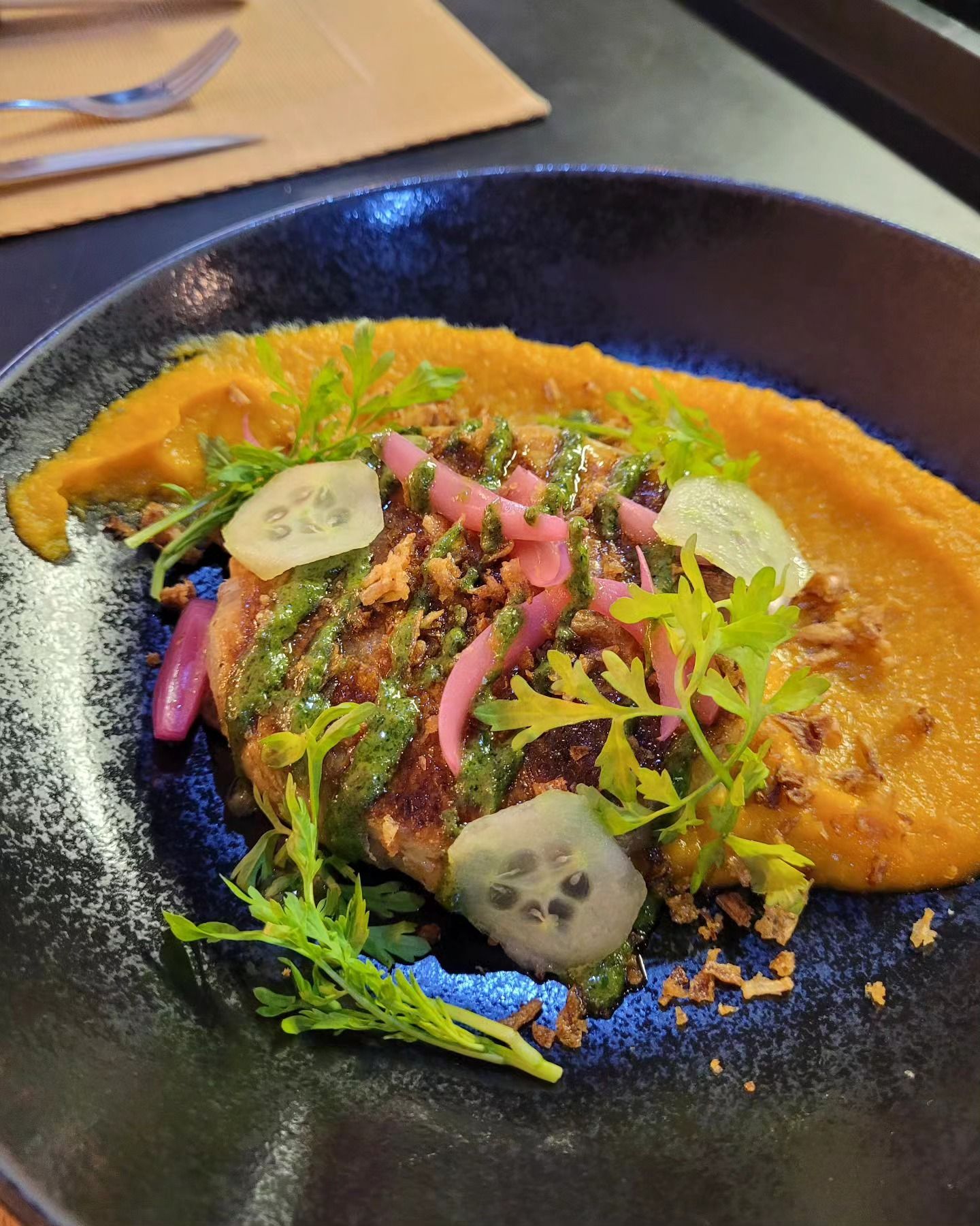 Assiette : Un steak haché doré avec de la purée de carottes, de la sauce verte, des oignons marinés et des légumes verts, sur une assiette bleu foncé.