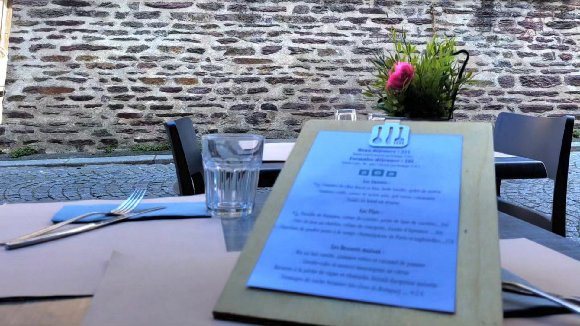 Table de restaurant dressée avec menu, verres et fleurs, adossée à un mur de briques.