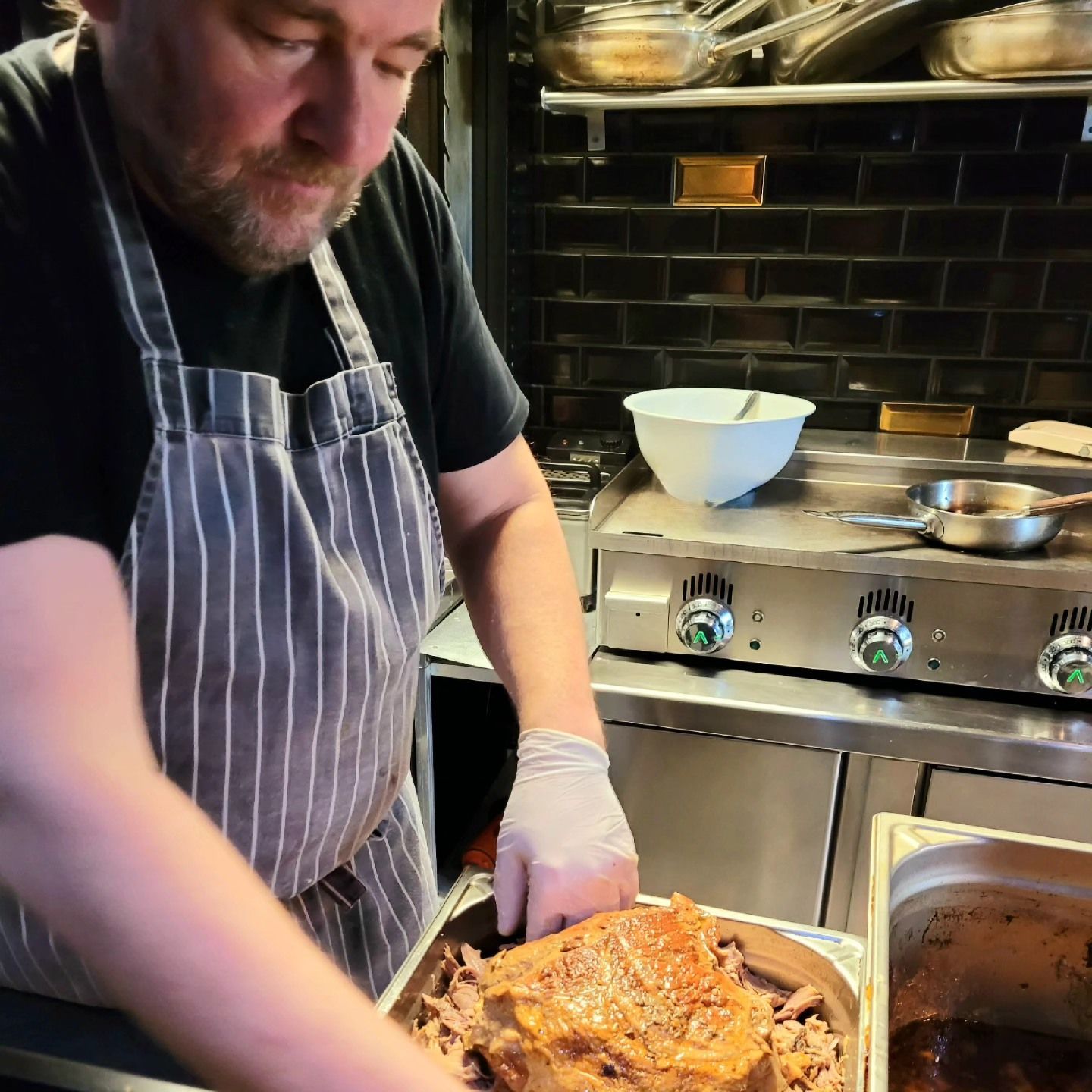 Un chef portant un tablier dans une cuisine professionnelle prépare des aliments, plongeant la main dans une poêle contenant de la viande rôtie.