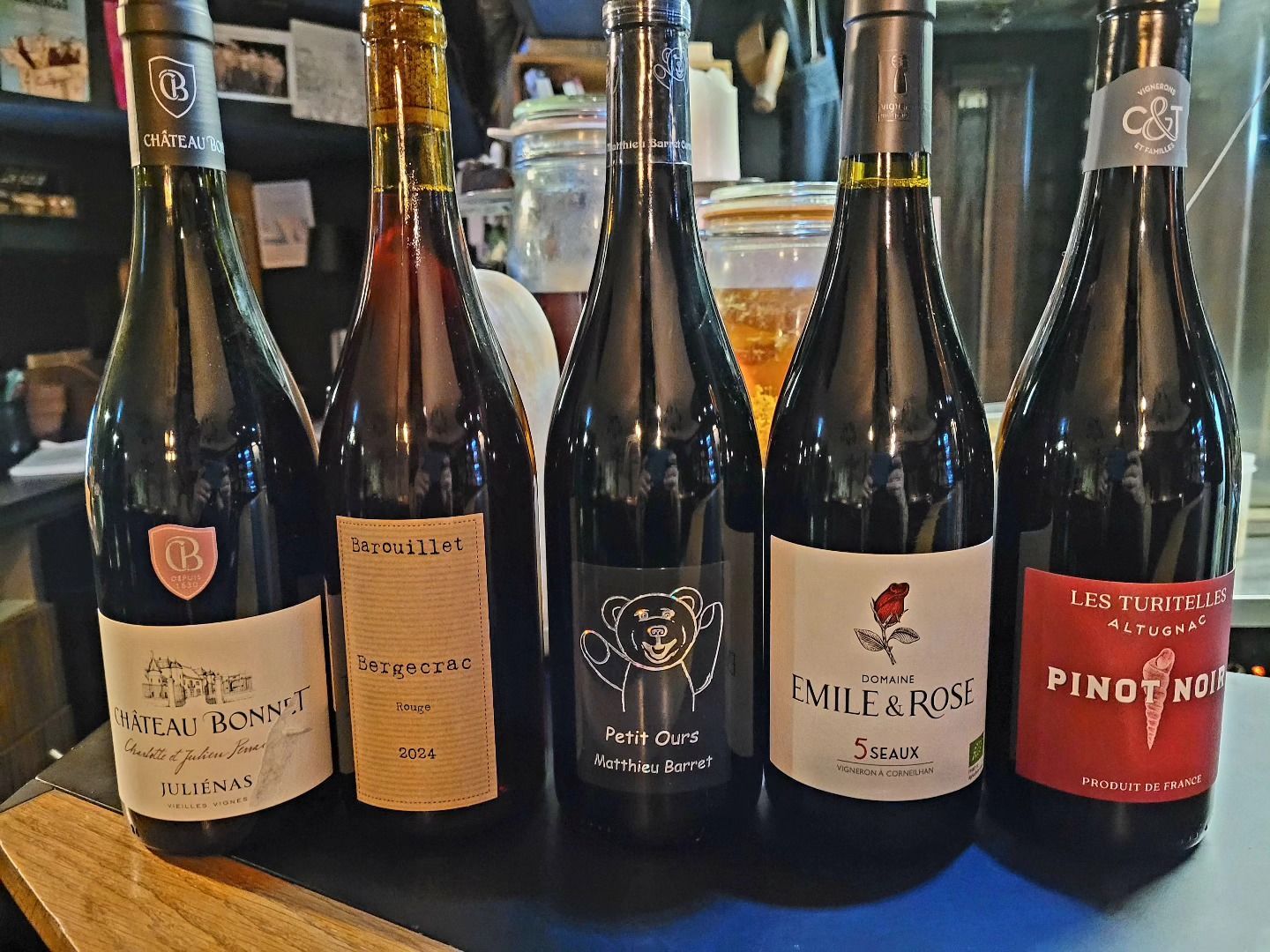 Cinq bouteilles de vin sur un bar, étiquettes variées, principalement des vins rouges, dans une ambiance sombre.