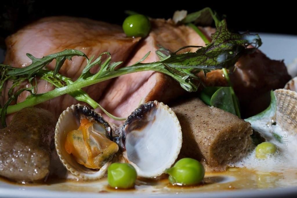 Gros plan sur un plat gastronomique : viande cuite, palourdes, petits pois et herbes, disposés sur une assiette avec de la sauce.